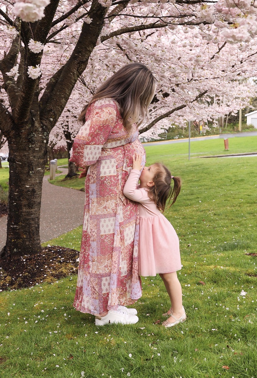 Mel with her daughter kissing her belly under cherry blossoms